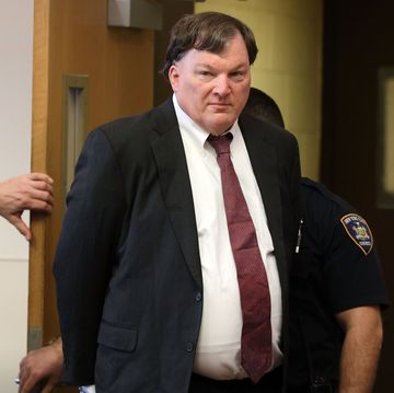 a man in a black suit jacket white collared shirt and red tie walks through a door as a police officer is behind him, the man's hands are behind his back and he looks to the left