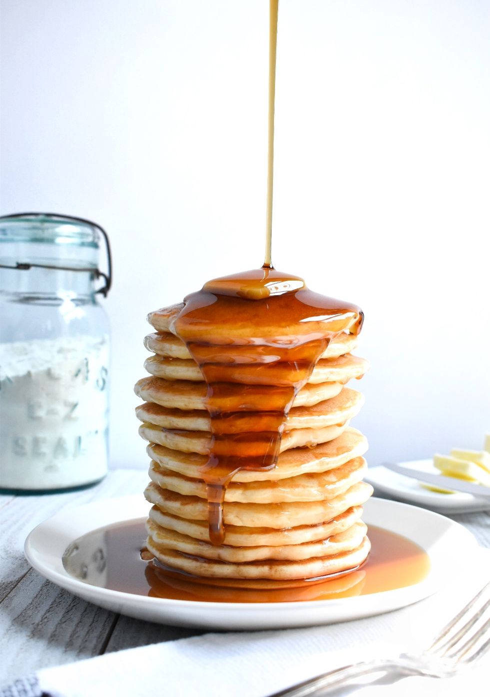 tall stack of pancakes with syrup being poured over them on a white plate alonside a fork, white napkin, a jar of flour and a dish of butter