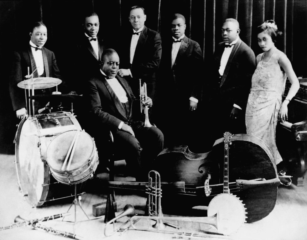 five men in tuxedos and one woman in a dress stand behind another man in a tuxedo who is seated, around them are several instruments including drums, a bass, horns, and a banjo