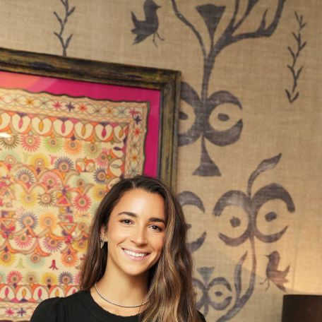 aly raisman smiles at the camera with one arm on her hip, she wears a black long sleeve top, gray skirt and silver chain necklace