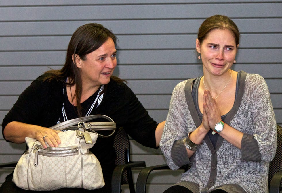 a woman sits in a chair next to amanda knox who is also seated, knox cries as her hands are held in front of her chest palms together
