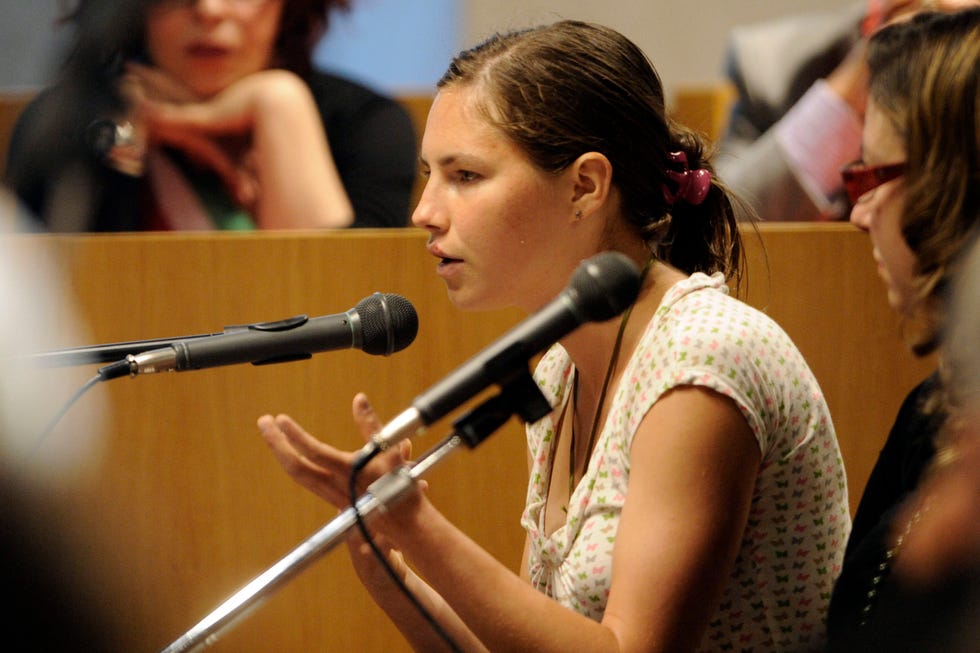 amanda knox speaks into a microphone that she sits behind and holds her hands in front of her, she wears a patterned short sleeve shirt with her hair pulled back