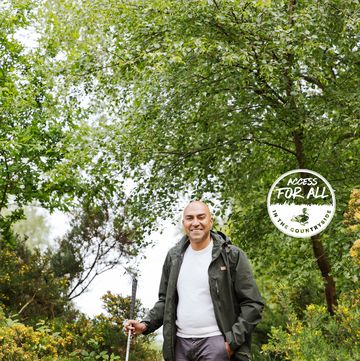 amar latif standing in the countryside surrounded by green trees he is wearing a rain jacket, white tshirt and grey trousers he looks happy and is smiling and the camera