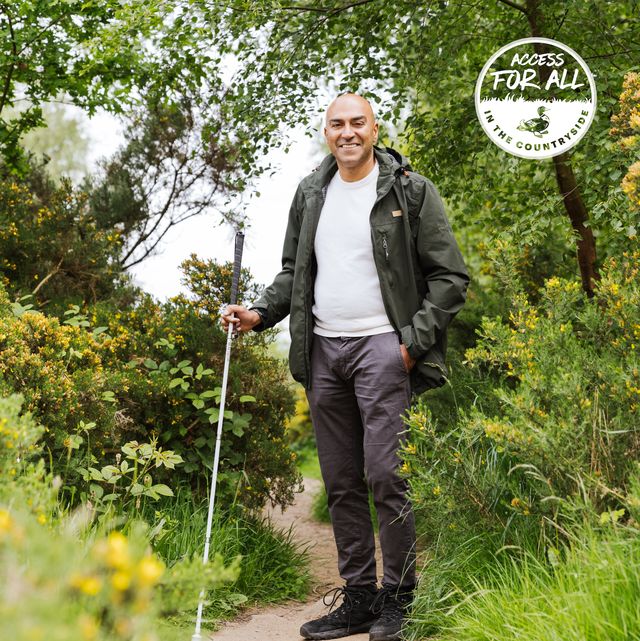 amar latif standing in the countryside surrounded by green trees he is wearing a rain jacket, white tshirt and grey trousers he looks happy and is smiling and the camera