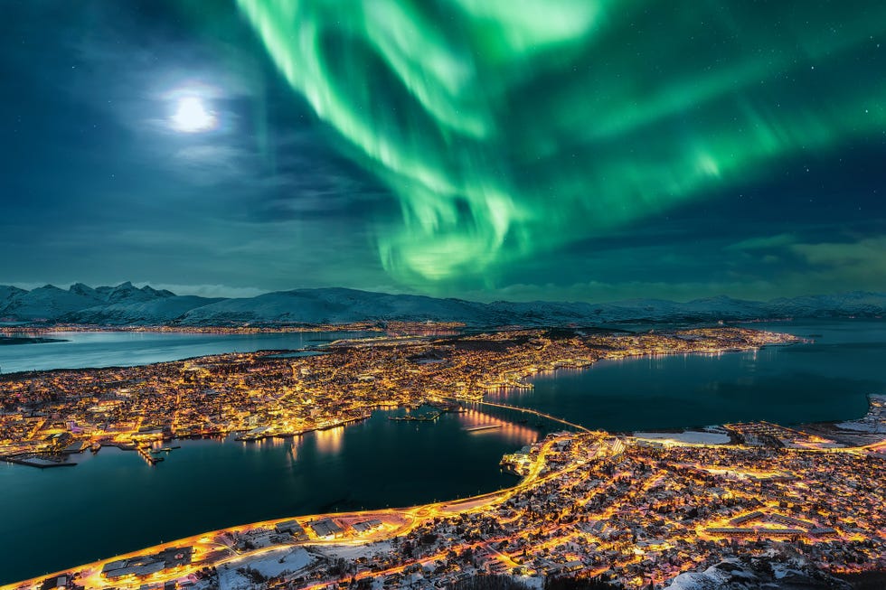 aurora borealis dancing over the city of tromso and full moon tromso urban skyline and winter night landscape with northern lights aurora polaris on a cold night, with starry sky, polar lights and snowcapped mountains on kvaloya island in the background cityscape with brightly street light of the arctic capital, northern norway