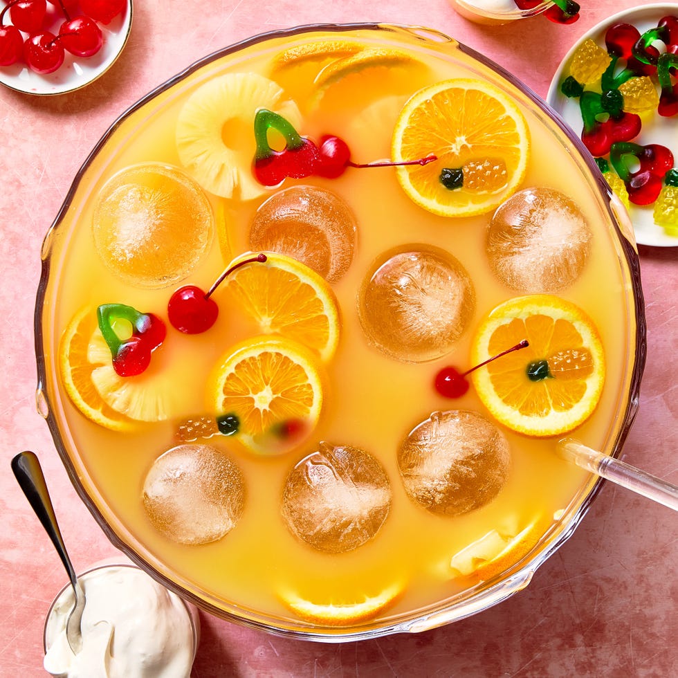 orange punch with round ice balls, sliced oranges, gummy candy, and maraschino cherries in a punch bowl with whipped cream on the side