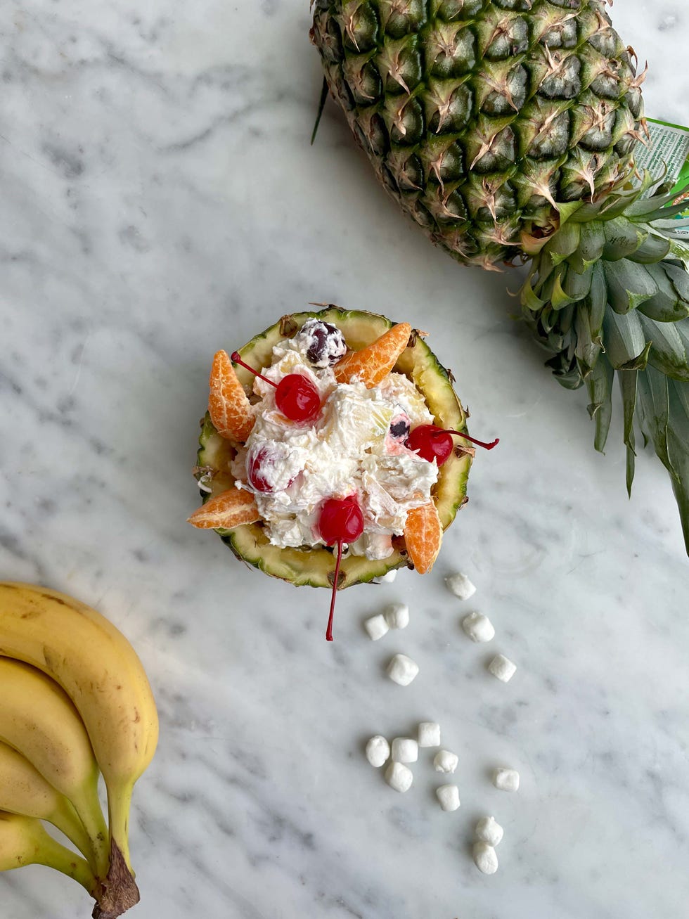 fruit salad served in a pineapple bowl with bananas and marshmallows nearby