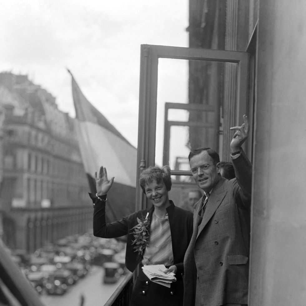 amelia earhart and george putnam wave and smile on a balcony overlooking a street, she holds a stack of papers and wears a sweater, blouse and skirt, he wears a suit