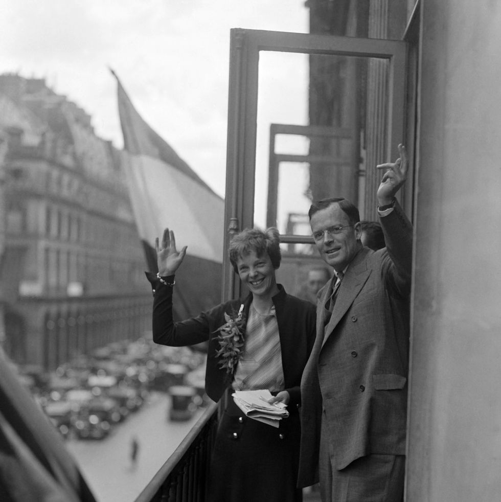 amelia earhart and george putnam wave and smile on a balcony overlooking a street, she holds a stack of papers and wears a sweater, blouse and skirt, he wears a suit