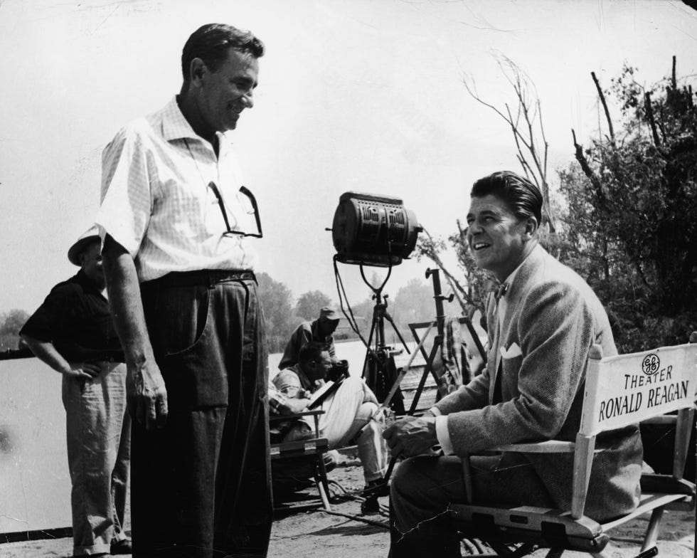 ronald reagan sits in a directors chair on a tv set and looks to the left toward a man standing there who looks at him