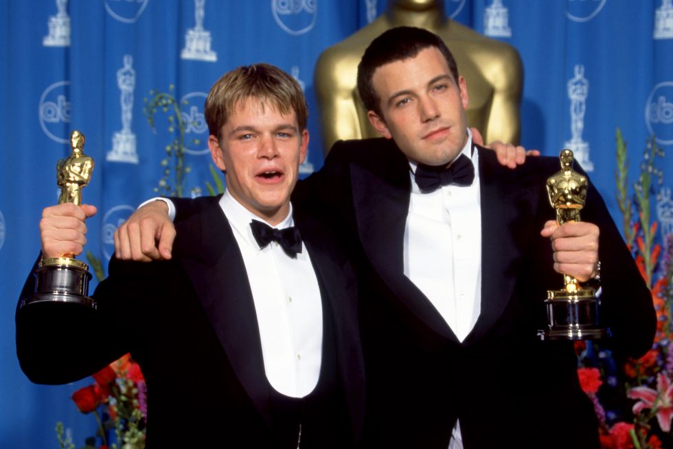 matt damon and ben affleck wearing tuxedos and holding oscar statuettes on a stage, standing in front of a blue curtain and large oscar statue