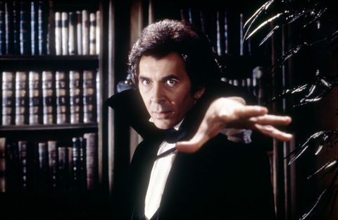 a promotional photo of frank langella in the film dracula, wearing a black and white suit, standing in front of bookshelves
