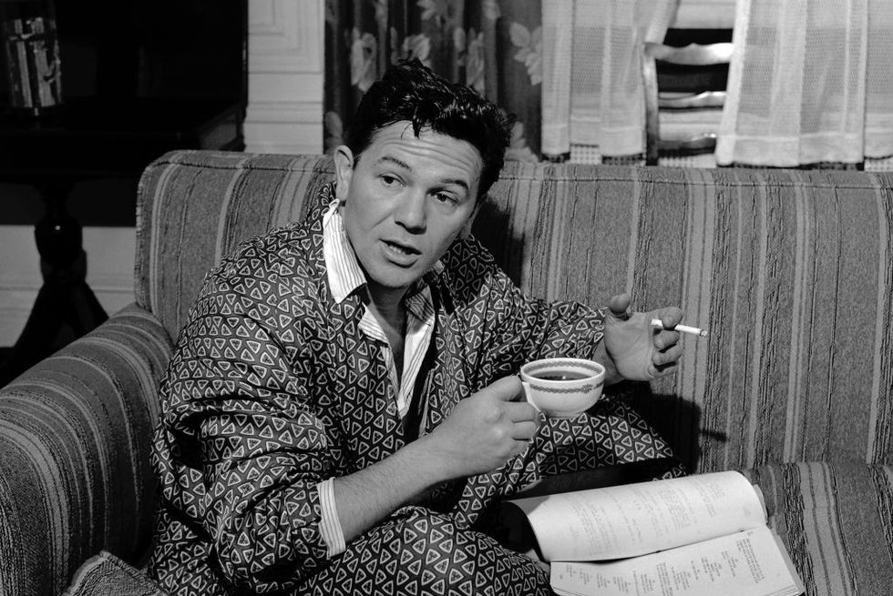 john garfield sits on coach wearing robe and holding a cigarette and cup of coffee