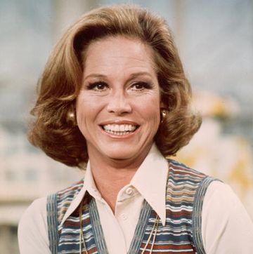 mary tyler moore smiles and looks past the camera, she is wearing a white collared shirt with a colorful striped vest on top, a long gold necklace, and gold hoop earrings
