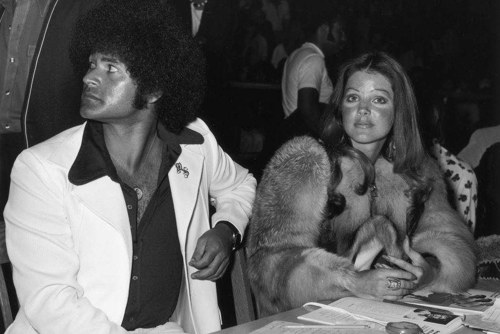 mike stone and priscilla presley sit at a table next to each other, he looks over his shoulder away from the camera and wears a white suit with a dark collared shirt, she looks at the camera and wears a fur jacket