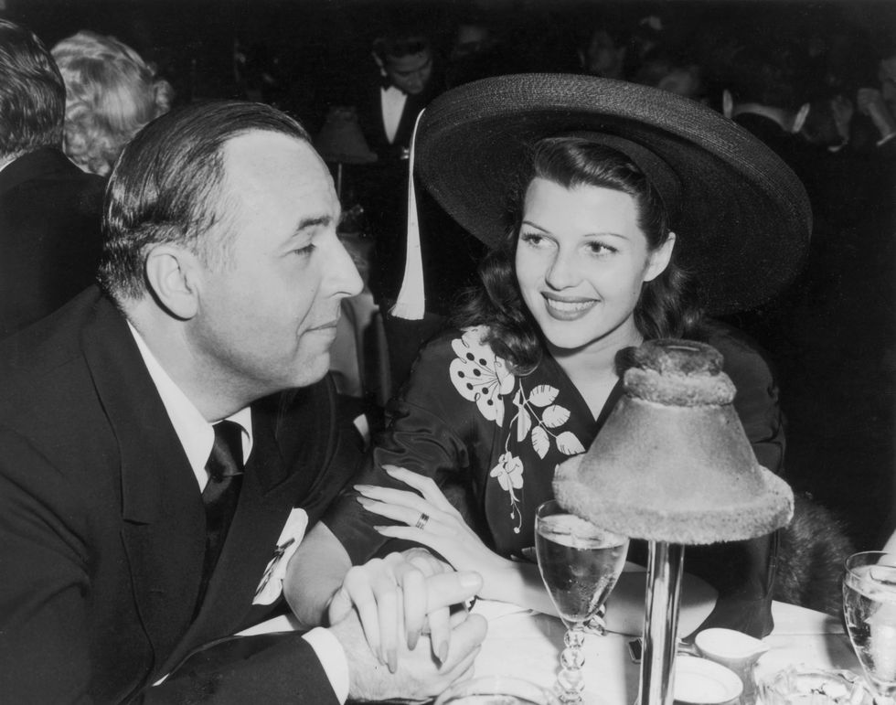 ed judson and rita hayworth sit at a restaurant table, he wears a suit, she wears a dress with a floral embroidered pattern and at hat, they hold hands and she smiles at him