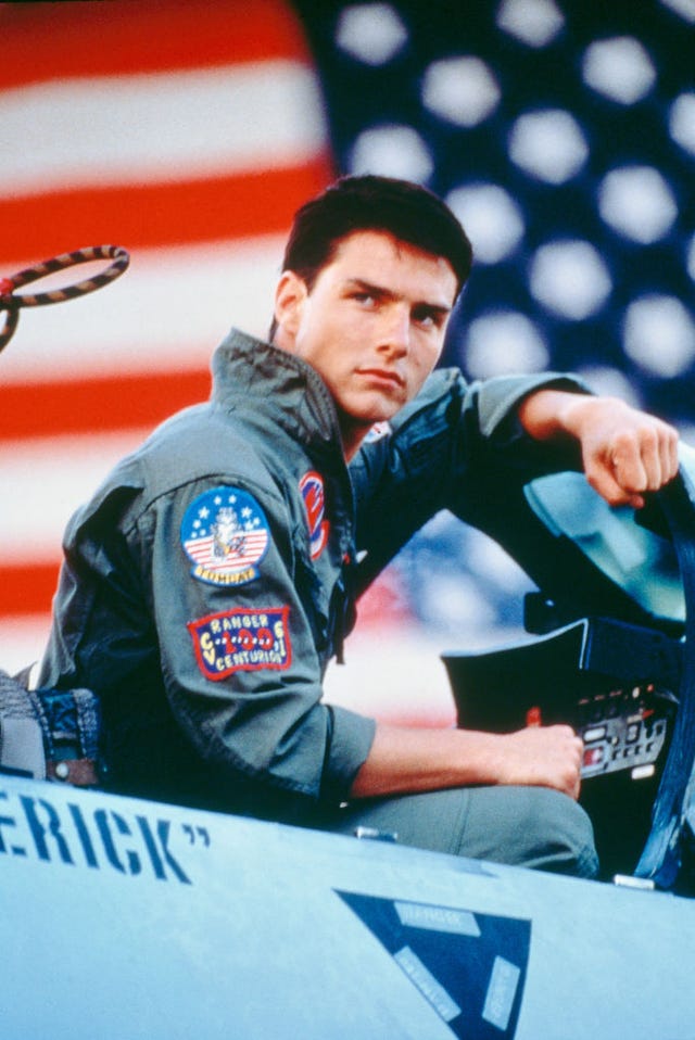 tom cruise poses in character for top gun, he sits in a fighter jet with the roof open and looks behind him, he wears a naval military uniform