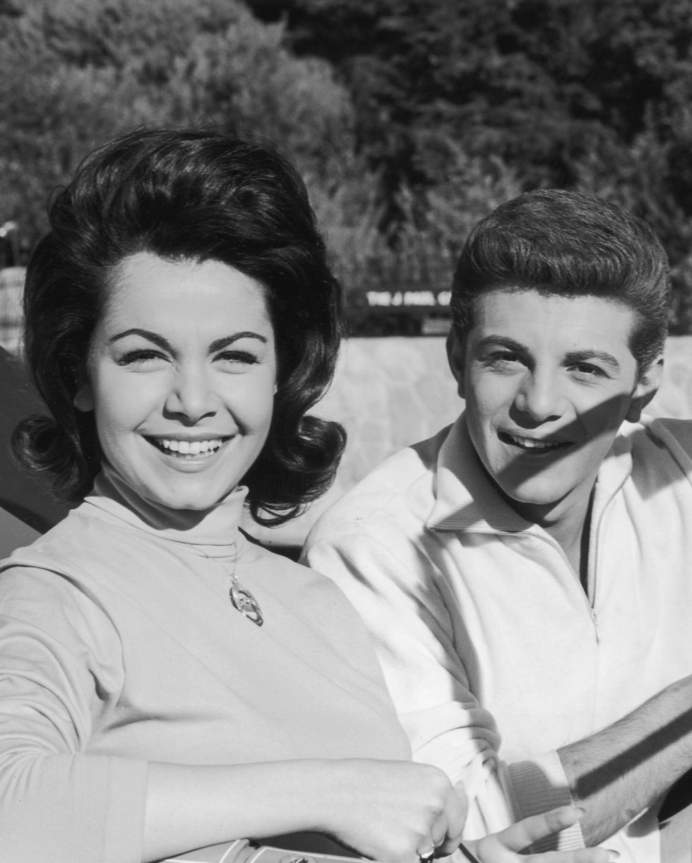 annette funicello and frankie avalon smile while sitting in the front seat of a convertible car, she wears a turtleneck long sleeve shirt with a necklace, he wears a quarter zip sweater