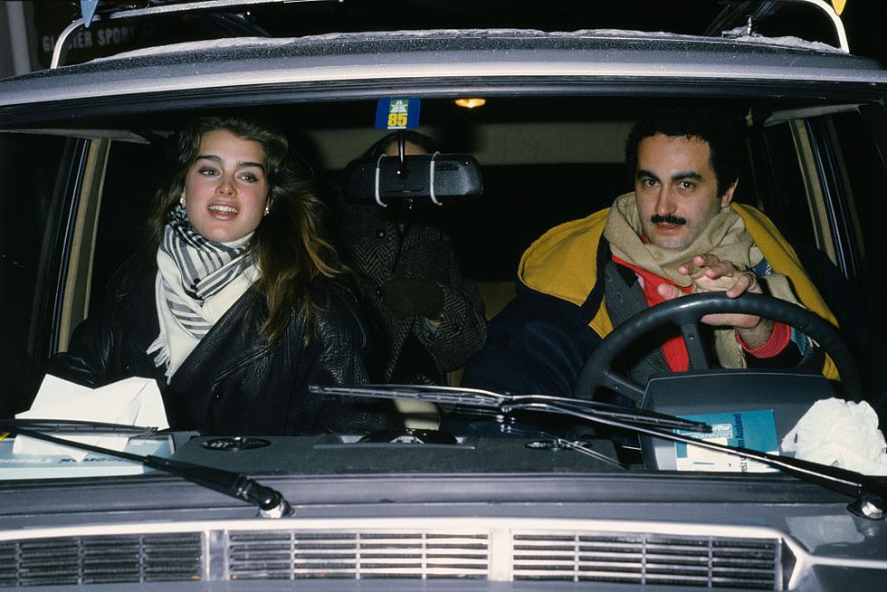 brooke shields and dodi fayed sit in a car wearing coats and scarfs, she is in the front passenger seat and he is in the drivers seat with one hand on the wheel