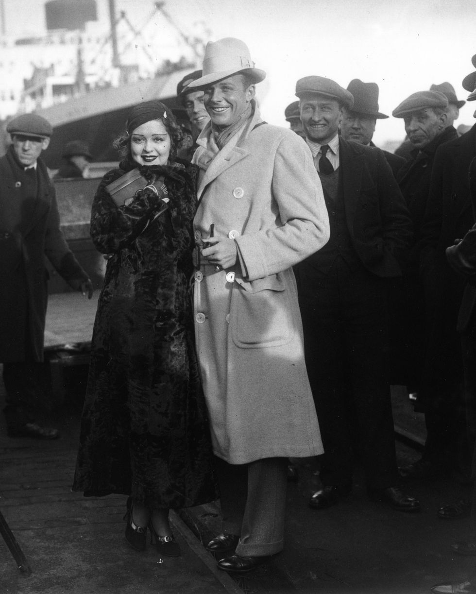 clara bow and rex bell stand in long coats outside and smile, both wear hats, a group of people stands behind, she holds a clutch to her body