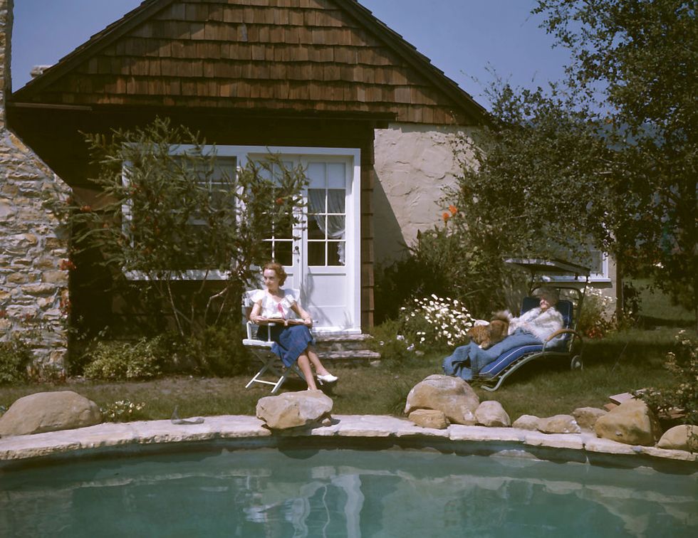 two women sit beside a pool in the backyard of a stove and wooden slate house on a sunny day
