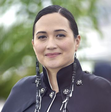 lily gladstone wearing a black outfit and native american jewelry, smiling directly at the camera