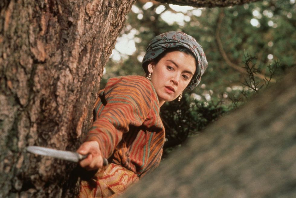a woman in costume holding a knife in defense while standing against a tree