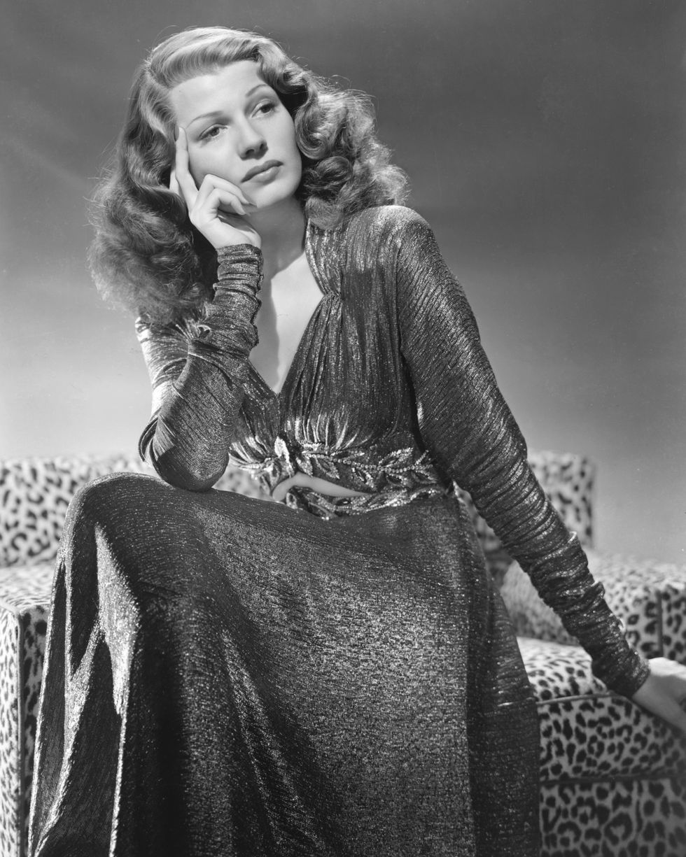 rita hayworth looks off into the distance with a sad face, she wears a shimmery long sleeve dress and sits on a cheetah print armchair