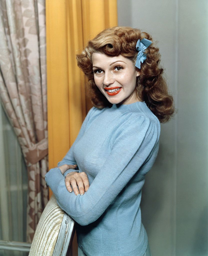 rita hayworth smiles as she looks over her shoulder, her arms are crossed and rest on a chair back, she wears a blue sweater and a blue hair ribbon