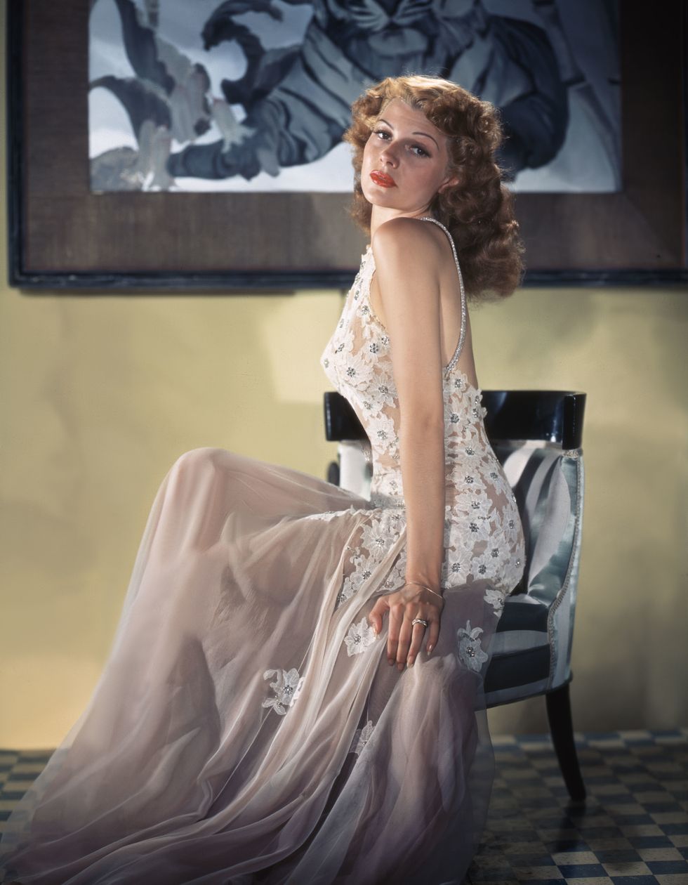 rita hayworth sits in a chair and looks over her shoulder at the camera, she wears a blush floor length dress with flower appliques
