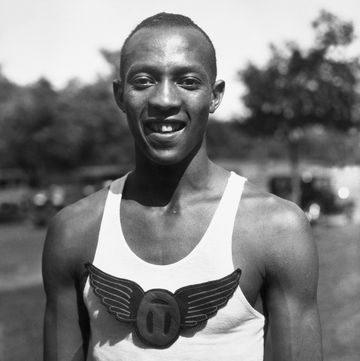 jesse owens smiles at the camera, he wears a light colored sleeveless shirt with a winged emblem in the center of the chest