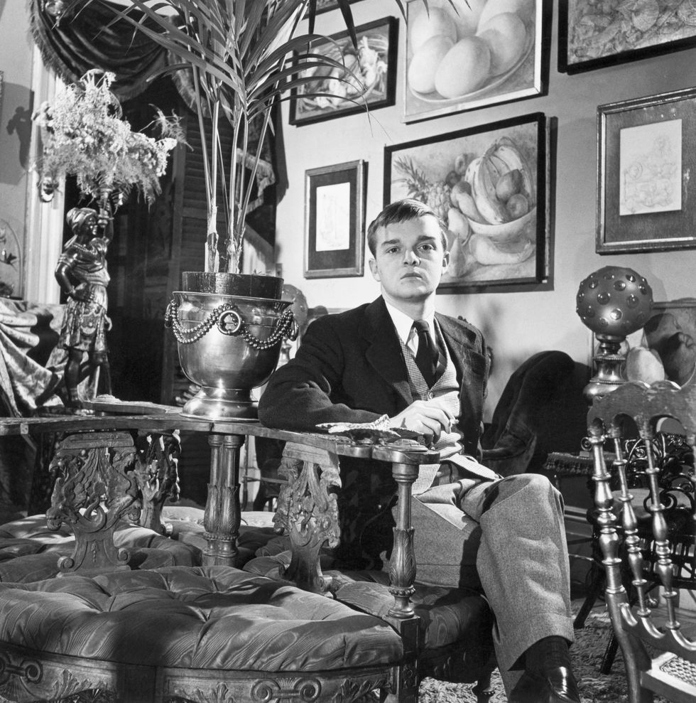 truman capote sits in a chair in a very decorated room, he wears a three piece suit with a tie and collared shirt