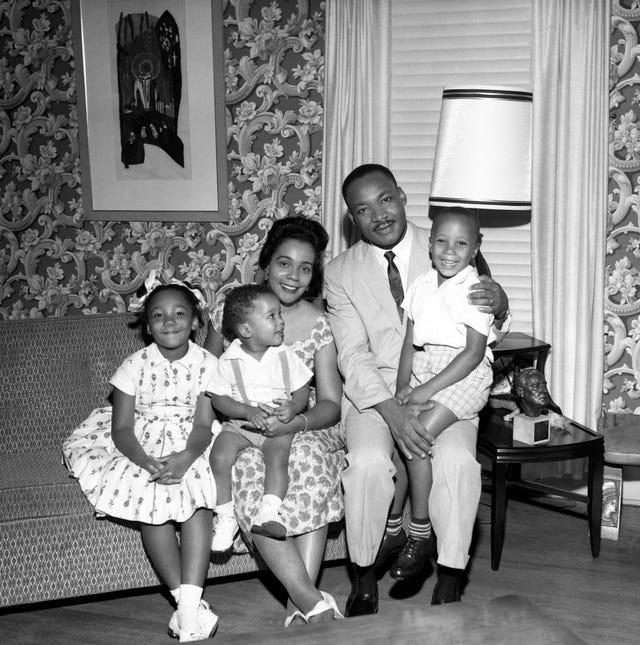 yolanda king, dexter king, coretta scott king, martin luther king jr, and martin luther king iii sit on a sofa and smile for a photo, they are in a room with patterned wallpaper and a window covered by blinds, a painting hangs on the wall and a side table is on the right with a table lamp