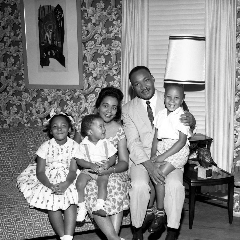 yolanda king, dexter king, coretta scott king, martin luther king jr, and martin luther king iii sit on a sofa and smile for a photo, they are in a room with patterned wallpaper and a window covered by blinds, a painting hangs on the wall and a side table is on the right with a table lamp