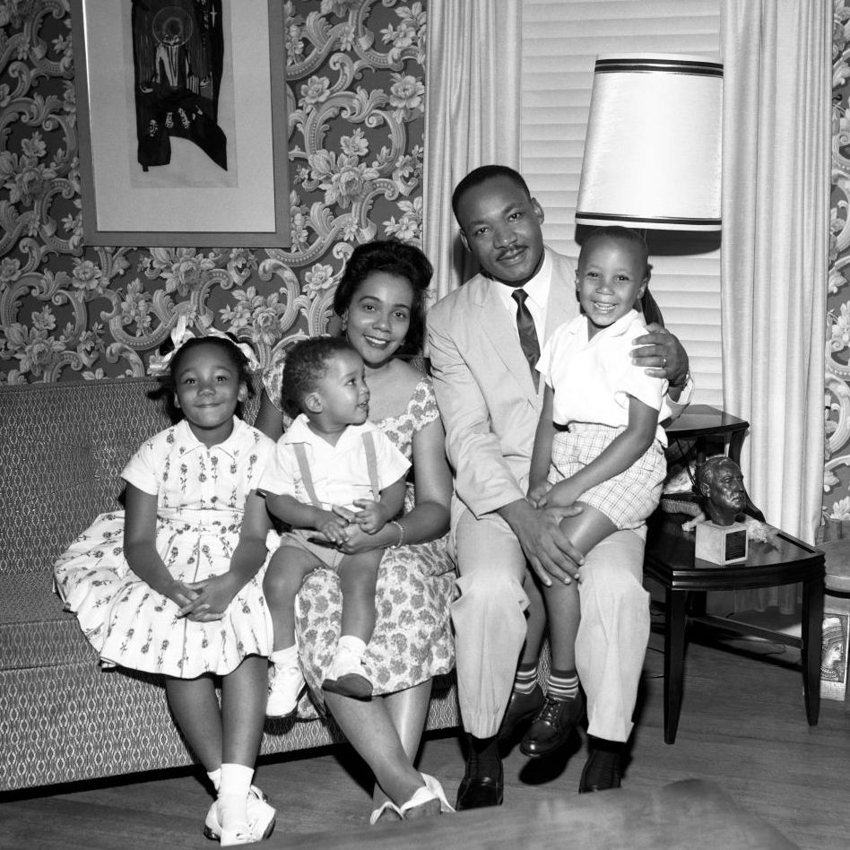 yolanda king, dexter king, coretta scott king, martin luther king jr, and martin luther king iii sit on a sofa and smile for a photo, they are in a room with patterned wallpaper and a window covered by blinds, a painting hangs on the wall and a side table is on the right with a table lamp