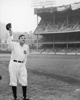 babe ruth holding up a baseball cap while wearing his yankees uniform inside a full baseball stadium