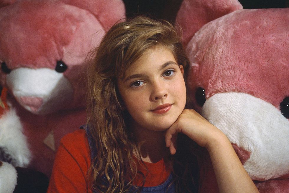 drew barrymore looks at the camera while resting her chin on one of her hands, she wears a red t shirt under a blue corduroy jumper, large pink teddy bears surround her