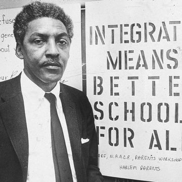bayard rustin looks at the camera while standing in front of antisegregation signs, he wears a suit jacket, collared shirt and tie and holds a cigarette in one hand