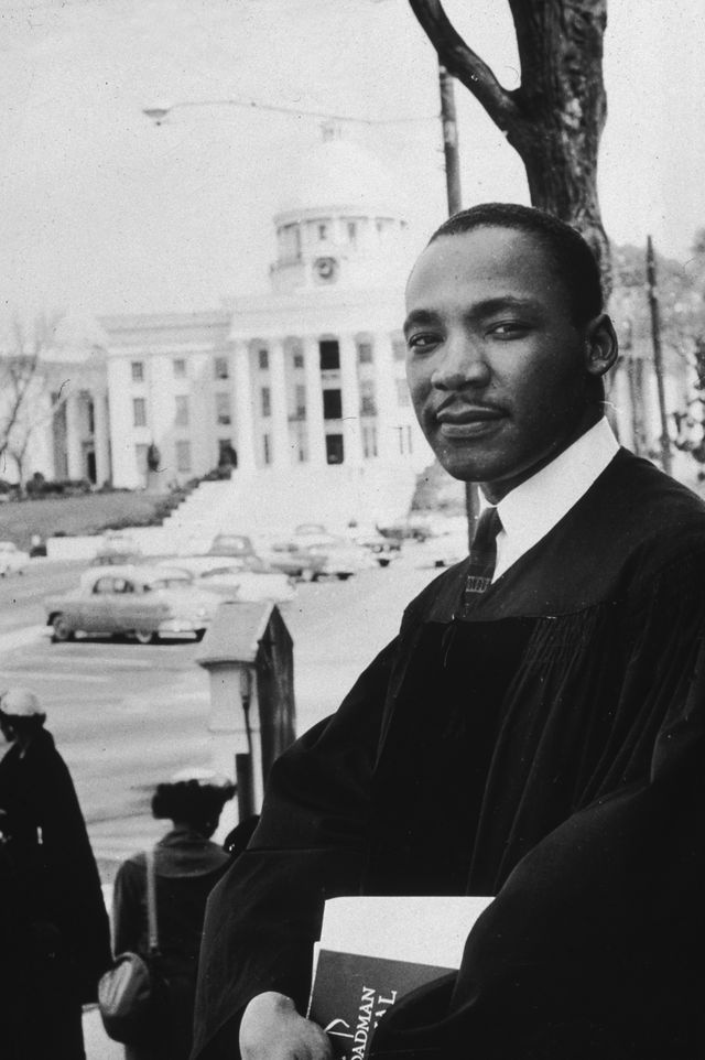 martin luther king jr looks at the camera while standing outside in a pastor robe over a collared shirt and tie, he holds papers in both hands in front of him, behind him is a street scene and a large white building