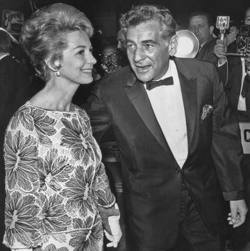 a black and white photo of felicia montealegre, wearing a floral dress, and leonardo bernstein, wearing a tuxedo, walking past reporters and photographers