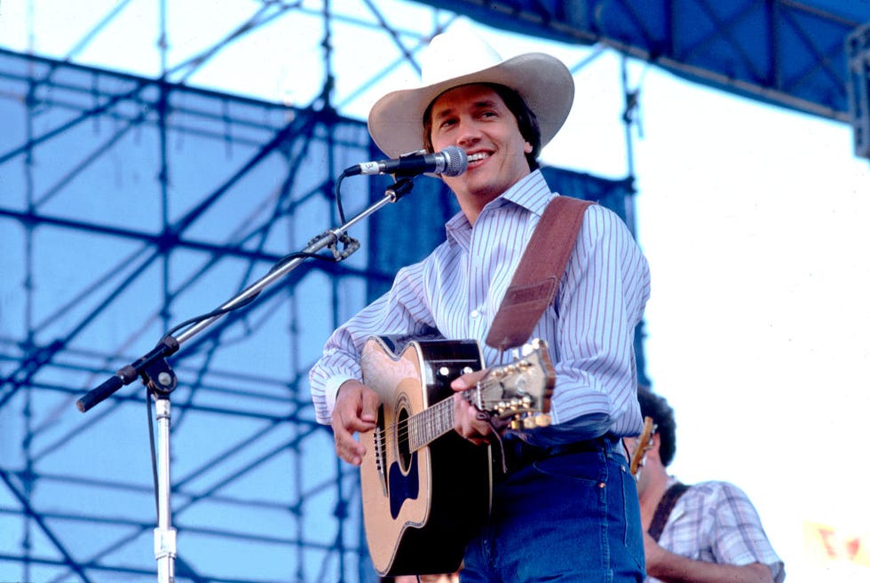 george strait performs on a stage outside, he wears a cowboy hat, collared shirt and jeans