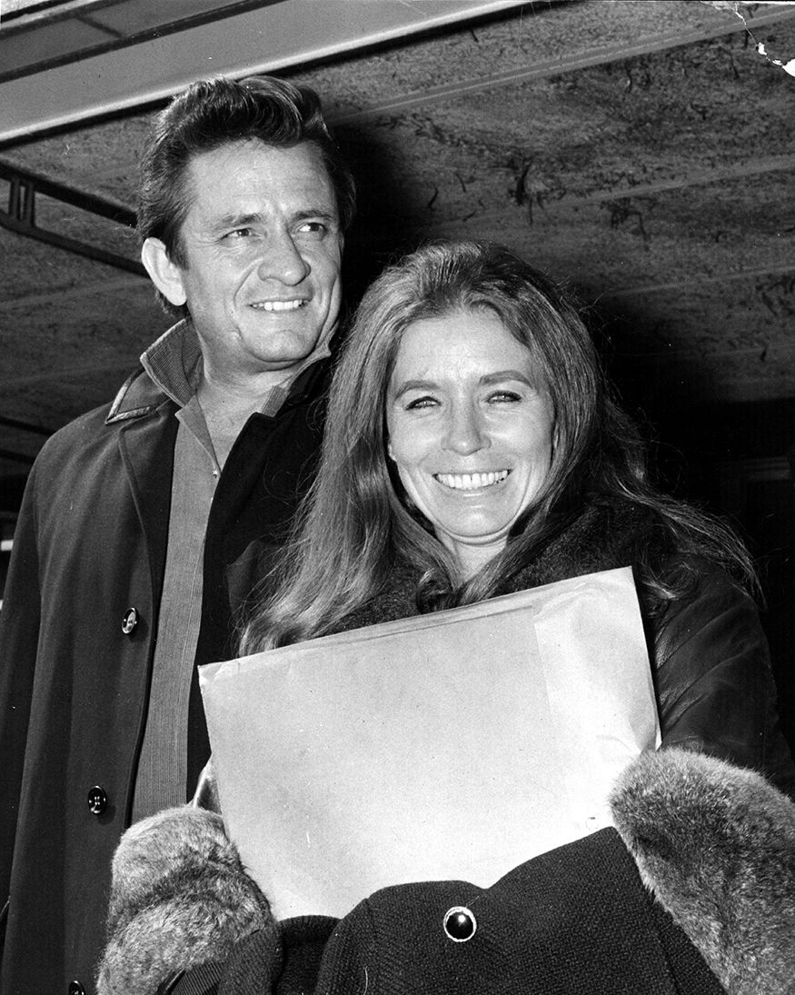johnny cash and june carter cash stand near one another and smile, he looks to the right, she looks straight at the camera and clutches a large envelope