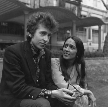 bob dylan sitting and holding a pair of glasses as joan baez looks over at him