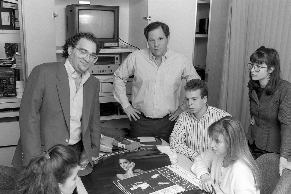 bob weinstein and harvey weinstein stand behind a table and look at the camera, three people are seated at the table and another woman stands to the right of the table, a tv and other equipment is in a cabinet behind them and movie posters are on the table