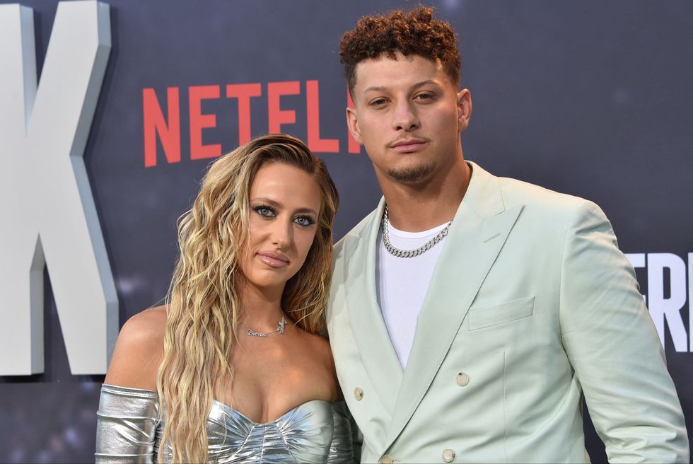 brittany mahomes and patrick mahomes pose for a photo in front of a dark gray background, she wears a strapless silver dress, he wears a baby blue suit jacket over a white shirt with a silver chain necklace