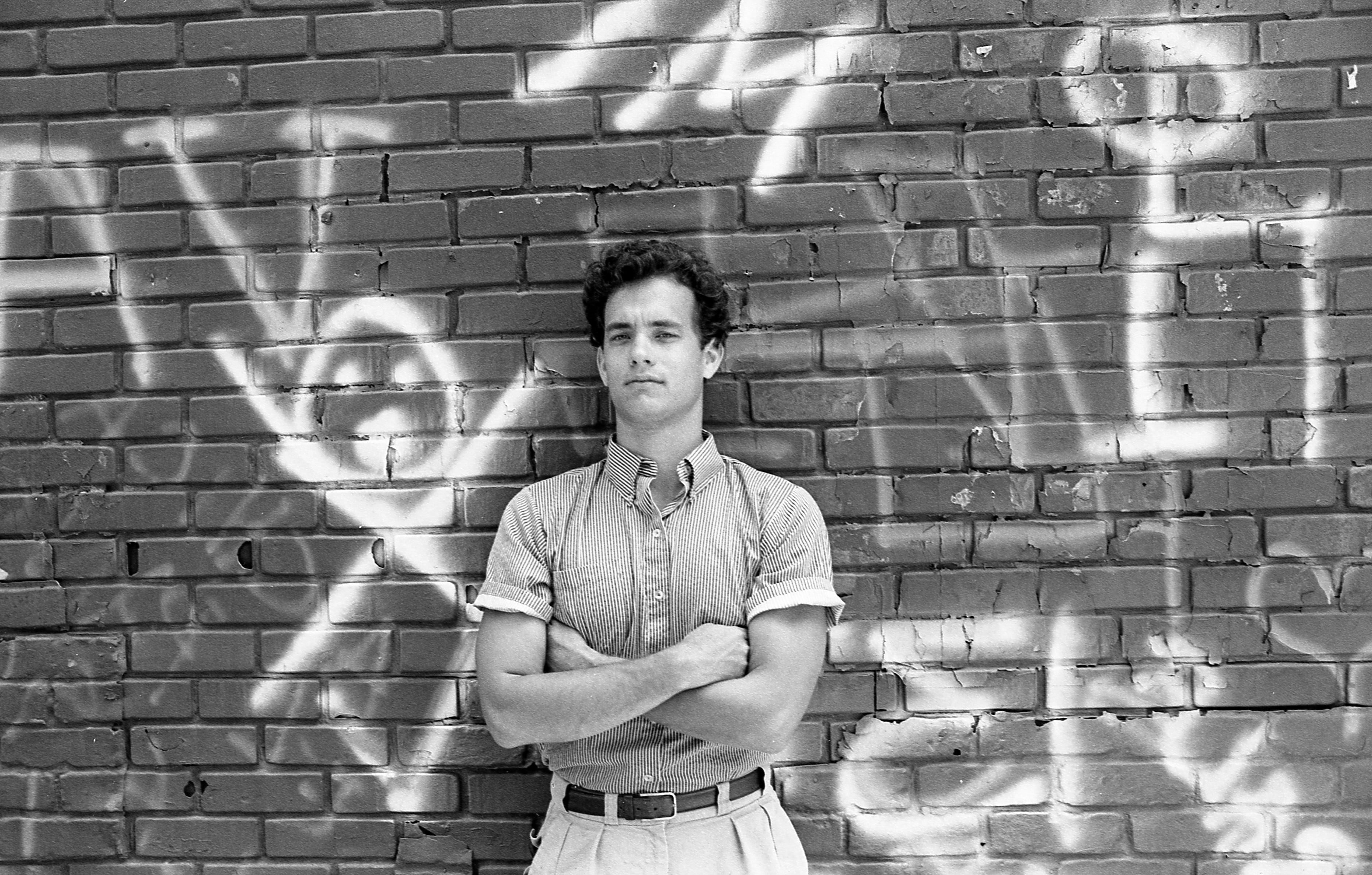 actor tom hanks leans his back against a brick wall with graffiti on it and looks at the camera, his arms are crossed on his chest, he wears a collared short sleeve shirt and light colored bottoms with a belt