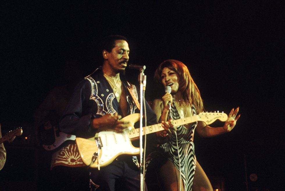 ike and tina turner performing on stage, ike stands in front of a microphone and plays guitar, tina stands on his right and sings into a microphone she is holding, ike wears a deep v cut black shirt with rhinestone details, tina wears a black and silver striped dress with high thigh slits