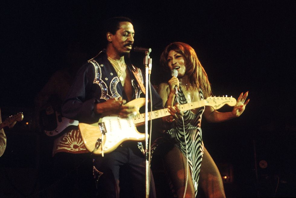 ike and tina turner performing on stage, ike stands in front of a microphone and plays guitar, tina stands on his right and sings into a microphone she is holding, ike wears a deep v cut black shirt with rhinestone details, tina wears a black and silver striped dress with high thigh slits