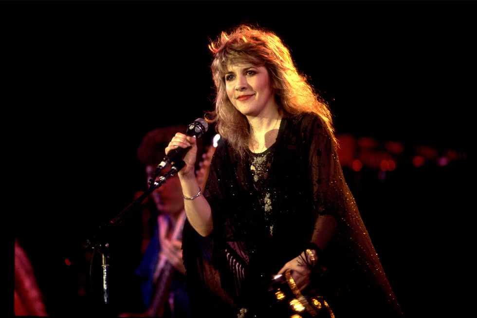 stevie nicks smiles while onstage, she grips a microphone on a stand in one hand and holds a tambourine in the other, her black glittery outfit glistens in the spotlight