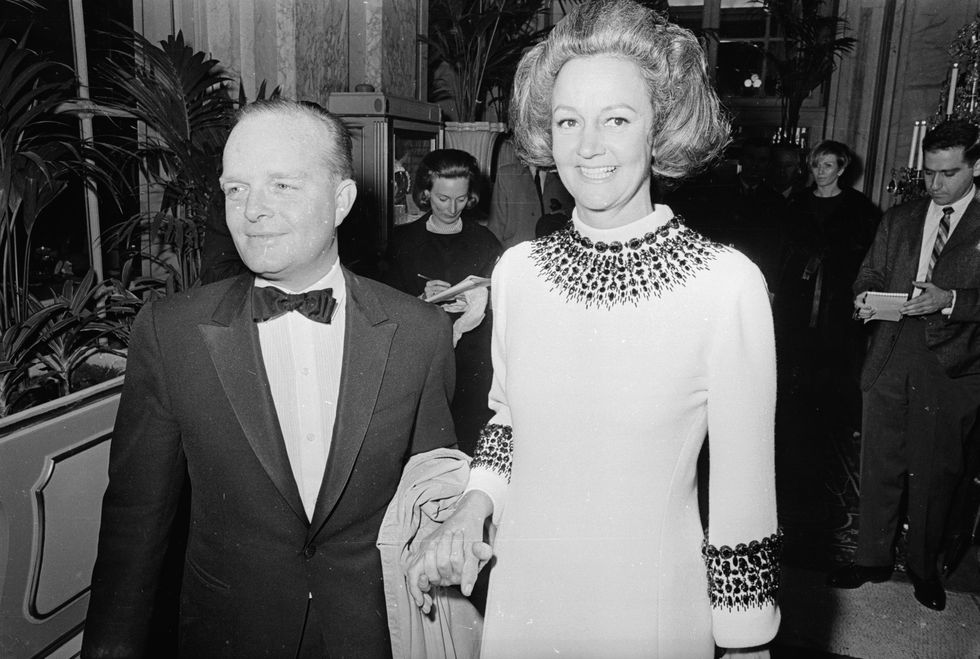 truman capote and katherine graham stand together in a hallway and smile, he wears a dark suit jacket with a bowtie, she wears a light colored dress with a pattern at the collar and cuffs